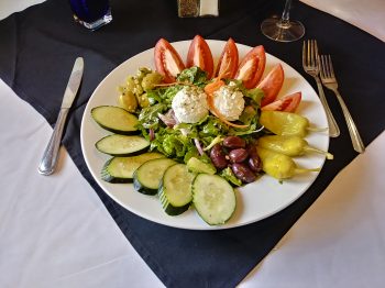 Photo of Jake and Telly's Family Sized Greek Salad on a white plate: Hand cut romaine lettuce, fresh tomato, cucumber, red onion, marinated olives, pepperoncini peppers, feta cheese topped with our house-made herbed vinaigrette dressing.