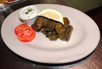 Vegetarian Appetizer: Jake and Telly's Dolmadakia (stuffed grape leaves) on a white plate. Imported Grape leaves stuffed with rice and onion, then cooked to perfection. Served with fresh tomatoes, lemon, and Tzatziki Sauce.