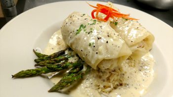 Jake and Telly's Specials - A close-up image of stuffed cabbage on top of asparagus and rice smothered with a white sauce and garnished with carrot and green parsley.
