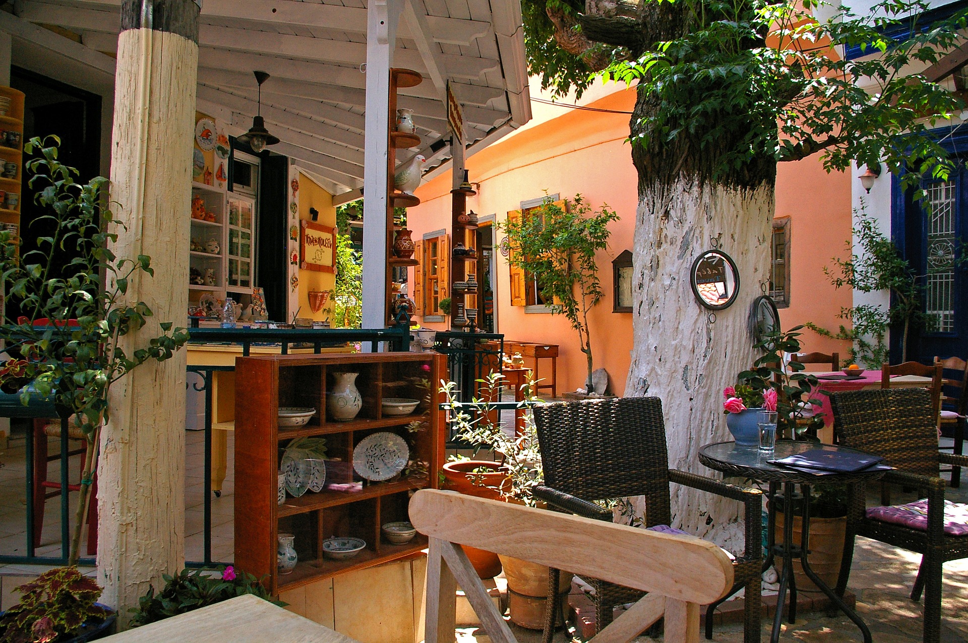 Jake and Telly's Catering: A photo of a covered, open-air patio on the Island of Samos. The patio is shaded by an open air roof and a large tree. Two tables are visible, along with three chairs, pottery on shelves and live potted trees and flowers. An orange wall is visible in the background.