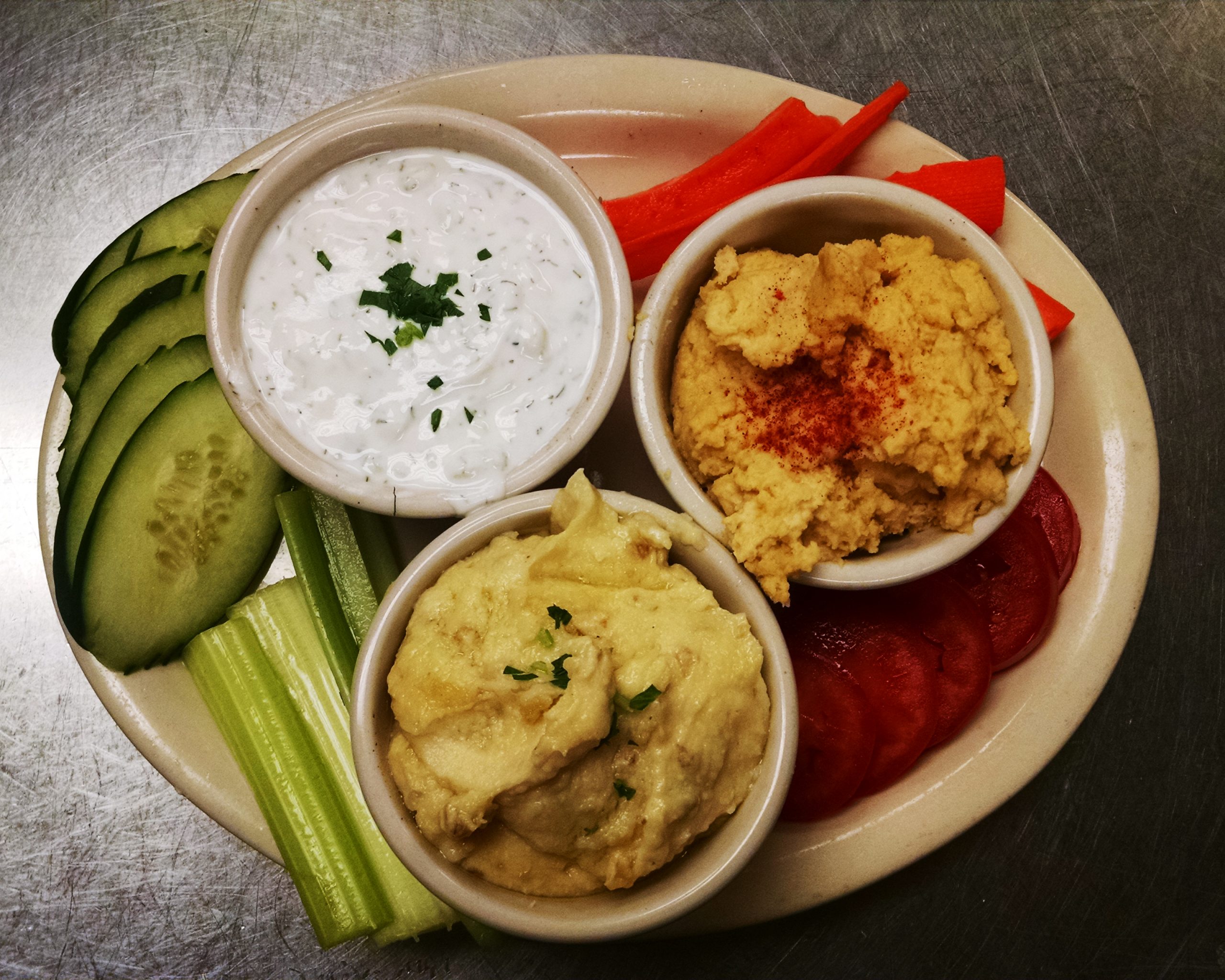 Appetizers: The Greek Dips. a Photo of our Hummus, Tzatziki and Skordalia on a white plate with fresh tomatoes, celery, carrots, and sliced cucumber.