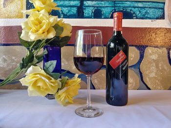 Nemea Red Wine from Greece: A photo of a bottle and glass of Nemea Red wine on a white tablecloth with yellow roses to their left. The wine glass is in the center, the bottle on the right. The collage is in front of a frescoed wall depicting a rock wall and a windowsill.