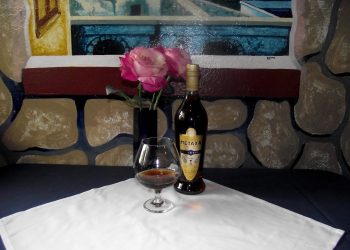 Metaxa: A photo of a brandy snifter with brown liquid setting on a white square tablecloth. A blue vase with pink roses is behind the brandy snifter to the left and a bottle of seven-star Metaxa is behind the brandy snifter to the right. the collage is in front of a frescoed wall depicting a rock wall and a windowsill.